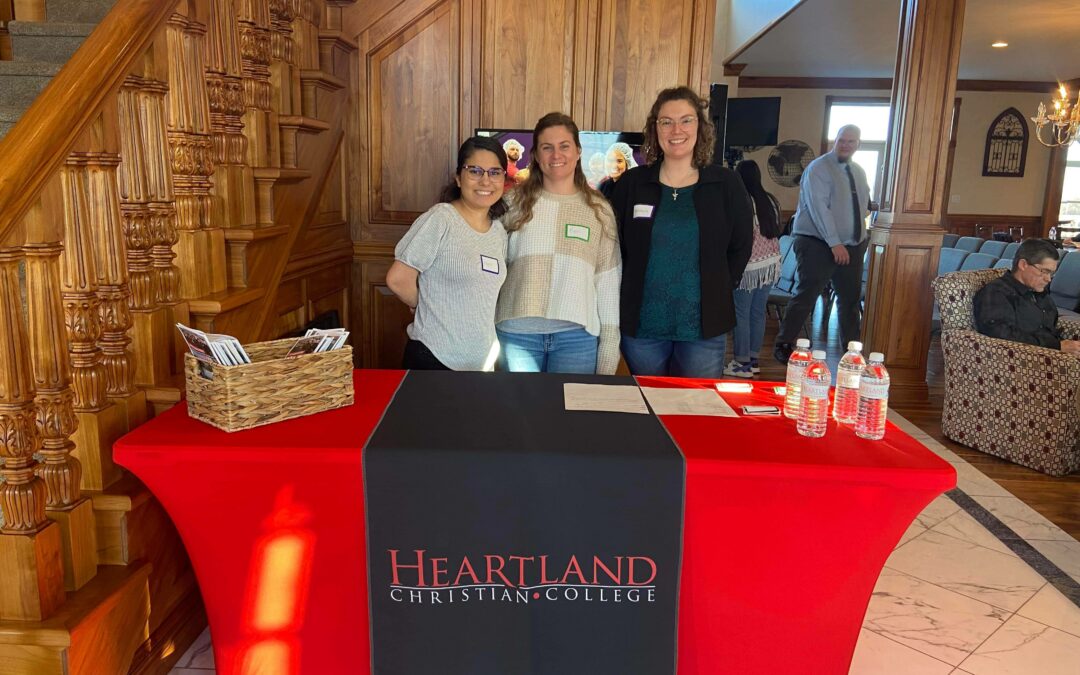 Three college girls standing behind a red and black registration table with the logo Heartland Christian College on the table. These students are responsible for registration during Preview Day 2024.